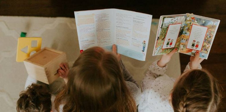 Presa zenital de dos infants asseguts a terra sobre una catifa, vistos d'esquena. Cadascun sosté un llibre obert: un sembla un llibre amb més text i l'altre un conte amb il·lustracions.
