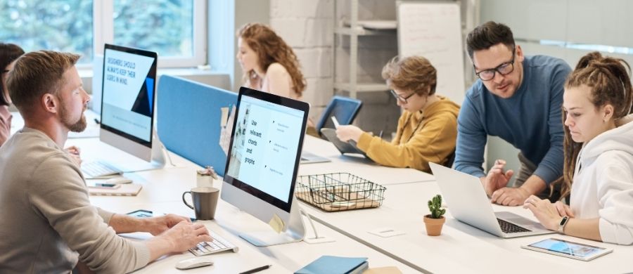 Sis persones treballen en un espai d’oficina o aula moderna. Estan assegudes davant d’ordinadors i portàtils. Un home ajuda una companya consultant la pantalla. Hi ha un ambient col·laboratiu.