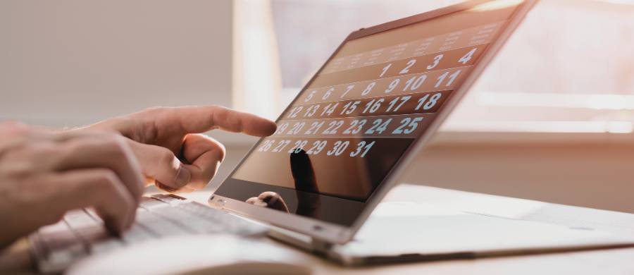 Una mano interactúa con una tableta en posición horizontal que muestra un calendario digital. Debajo hay un teclado y documentos sobre la mesa.