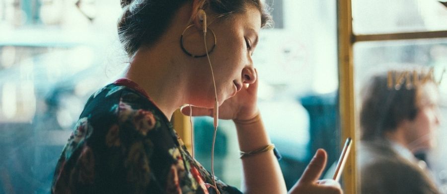  Dona jove asseguda al costat d’una finestra, escoltant amb auriculars petits mentre mira el telèfon mòbil. Porta arracades grans i una brusa amb estampat floral. L’escena és tranquil·la i amb llum natural suau.