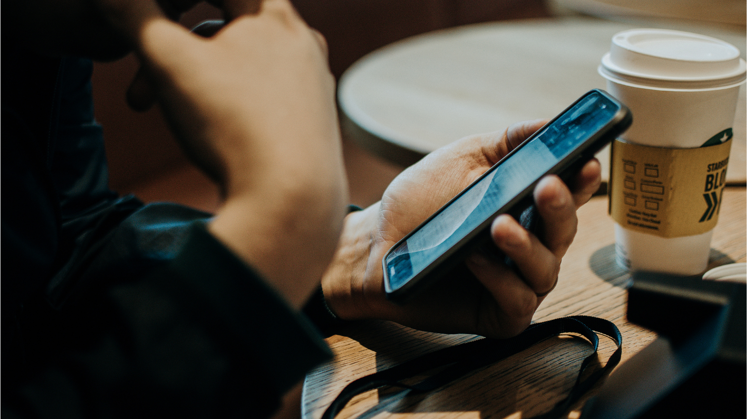 Un primer pla de les mans d'una persona que sosté un telèfon intel·ligent mentre seu a una taula de fusta. Al costat, hi ha un got de cafè per emportar.
