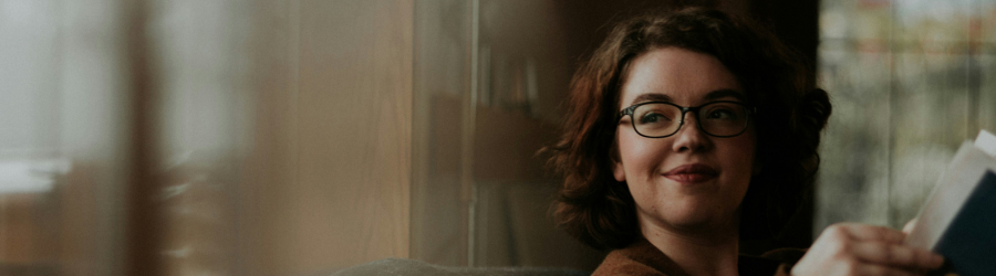 A woman with glasses and curly hair gently smiles while holding a book in a warm and cozy setting. A woman with glasses and curly hair gently smiles while holding a book in a warm and cozy setting.