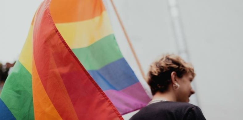 Persona de perfil con el pelo corto y pendientes, junto a una bandera arcoíris, símbolo del colectivo LGTBI+, ondeando en un entorno urbano desenfocado.