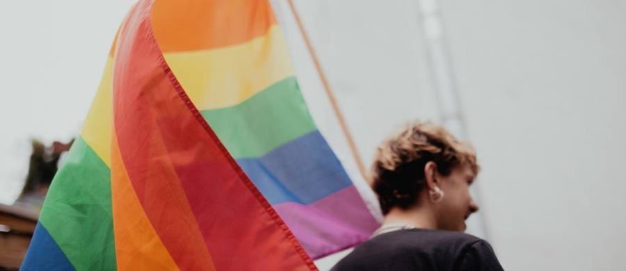 Persona de perfil con el pelo corto y pendientes, junto a una bandera arcoíris, símbolo del colectivo LGTBI+, ondeando en un entorno urbano desenfocado. Persona de perfil con el pelo corto y pendientes, junto a una bandera arcoíris, símbolo del colectivo LGTBI+, ondeando en un entorno urbano desenfocado.