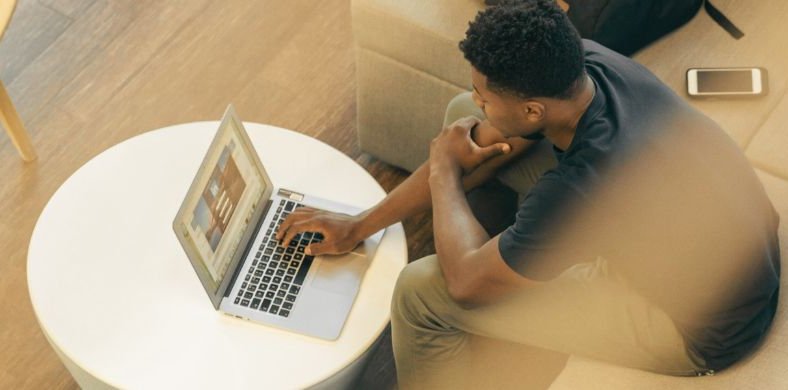 A young man is sitting on a sofa, working on a laptop placed on a round white table. He has a focused posture and a phone next to him.