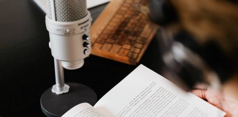 Persona con auriculares leyendo un libro abierto frente a un micrófono de pie sobre una mesa negra. También se ve parcialmente un teclado retro de madera.