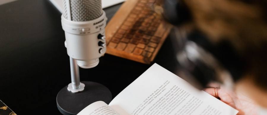 Persona con auriculares leyendo un libro abierto frente a un micrófono de pie sobre una mesa negra. También se ve parcialmente un teclado retro de madera.