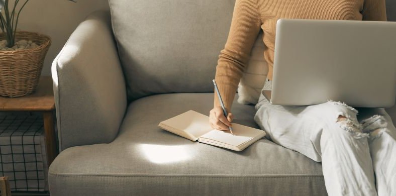 A woman sitting on the sofa with the computer in her legs while writing with a pencil in a notebook