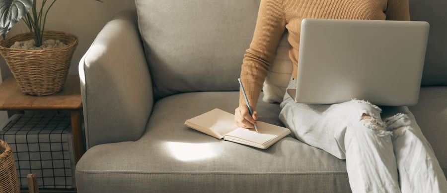 A woman sitting on the sofa with the computer in her legs while writing with a pencil in a notebook