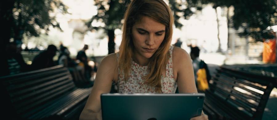 Una mujer joven con el pelo castaño y largo trabaja concentrada con una tableta digital mientras está sentada en un banco de madera en un parque. Una mujer joven con el pelo castaño y largo trabaja concentrada con una tableta digital mientras está sentada en un banco de madera en un parque.