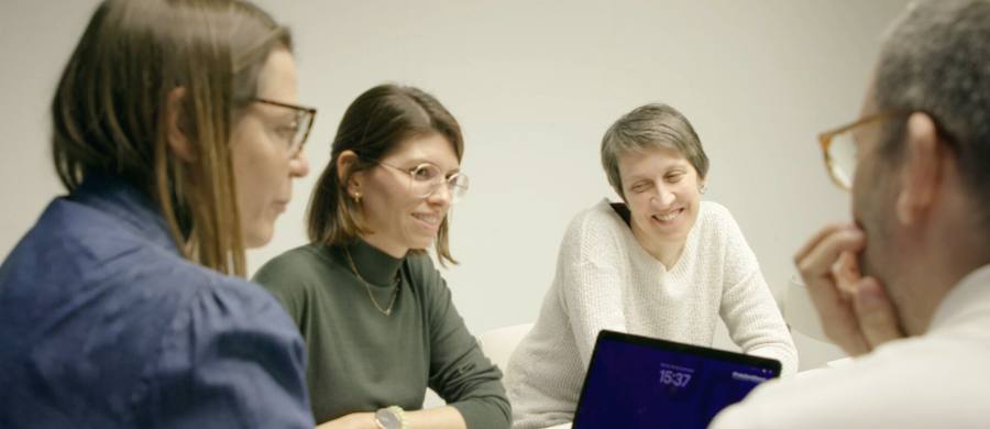 Captura del nou vídeo de la Biblioteca. Quatre persones en una reunió de treball, assegudes al voltant d’una taula. Dues dones amb ulleres escolten atentes, mentre una altra somriu. Un home amb ulleres reflexiona amb la mà a la barbeta.  Ambient distès i col·laboratiu.