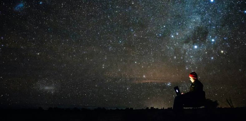 Una persona amb un gorro seu a l'aire lliure de nit, utilitzant un ordinador portàtil la pantalla del qual li il·lumina la cara, sota un vast cel nocturn ple d'estrelles i la Via Láctea.