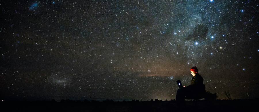 Una persona amb un gorro seu a l'aire lliure de nit, utilitzant un ordinador portàtil la pantalla del qual li il·lumina la cara, sota un vast cel nocturn ple d'estrelles i la Via Láctea. Una persona amb un gorro seu a l'aire lliure de nit, utilitzant un ordinador portàtil la pantalla del qual li il·lumina la cara, sota un vast cel nocturn ple d'estrelles i la Via Láctea.
