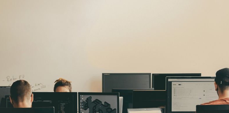  A group of people is working in front of computers in a room with a light-colored wall and some writings on the whiteboard.