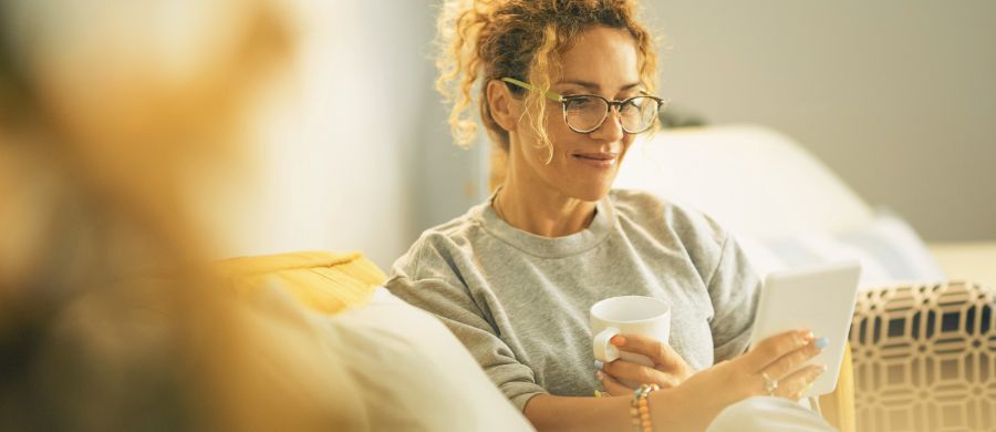 Una mujer sentada en el sofá leyendo un libro digital en la tableta con una taza en la mano.