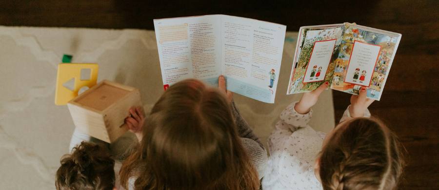 Presa zenital de dos infants asseguts a terra sobre una catifa, vistos d'esquena. Cadascun sosté un llibre obert: un sembla un llibre amb més text i l'altre un conte amb il·lustracions.