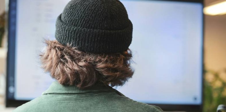 The image shows a person with wavy hair, wearing a dark beanie and a green jacket, sitting in front of a large screen in a modern work environment.