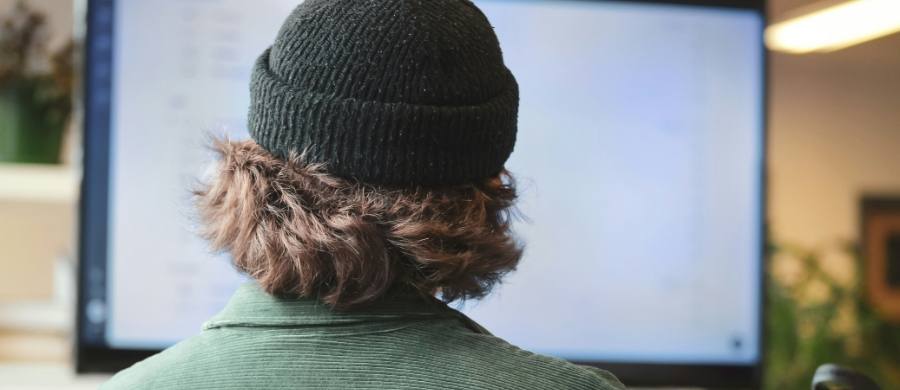 La imagen muestra a una persona de cabello ondulado con un gorro oscuro y chaqueta verde, sentada frente a una gran pantalla en un entorno de trabajo moderno.