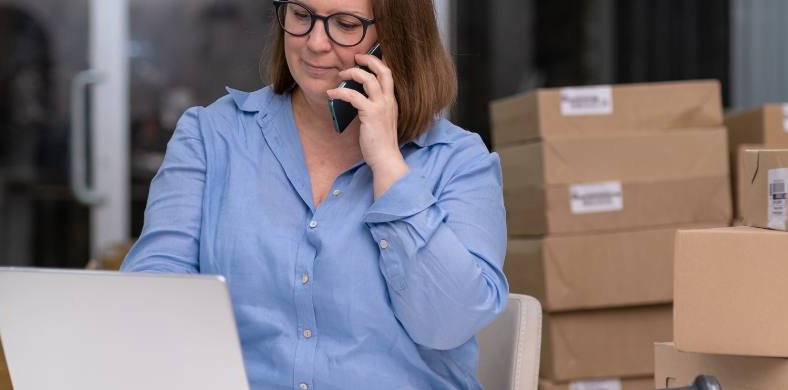 A woman talking on the phone with a computer in front and packages in the background