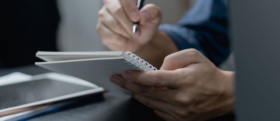 Primer plano de una persona escribiendo en una libreta de anillas con una mano, mientras sostiene la libreta con la otra. También se ve parcialmente una tableta digital sobre la mesa. Primer plano de una persona escribiendo en una libreta de anillas con una mano, mientras sostiene la libreta con la otra. También se ve parcialmente una tableta digital sobre la mesa.