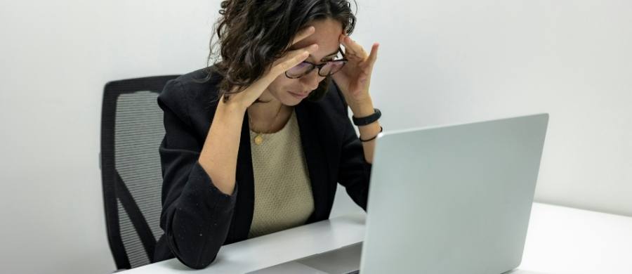 Mujer joven sentada frente a un portátil, con expresión de cansancio o estrés, tocándose la frente con las manos. Mujer joven sentada frente a un portátil, con expresión de cansancio o estrés, tocándose la frente con las manos.