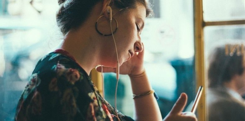 Mujer joven sentada junto a una ventana, escuchando con auriculares pequeños mientras mira su teléfono móvil. Lleva pendientes grandes y una blusa con estampado floral. La escena es tranquila y con luz natural suave.
