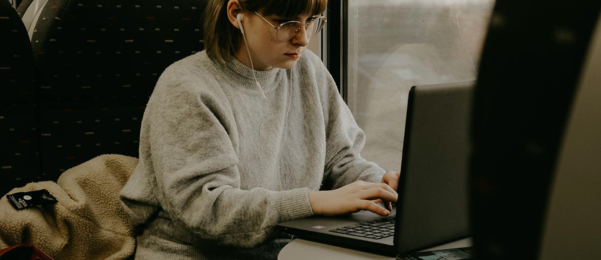 A girl sitting on a train with a pair of headphones and writing on a laptop