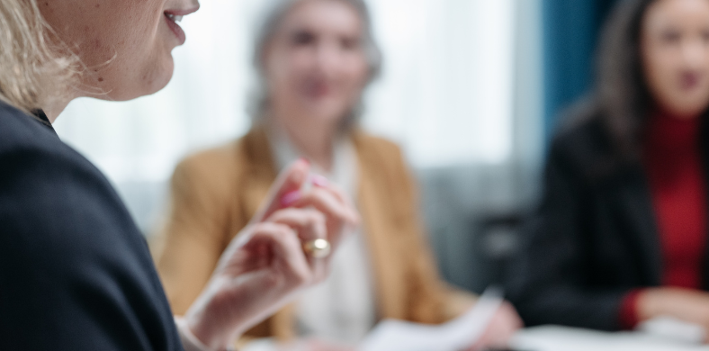 Un primer plano de una mujer hablando. En el fondo desenfocado, otras dos mujeres están sentadas en lo que parece una reunión de trabajo.