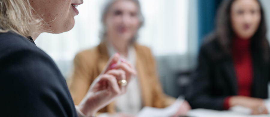 Un primer plano de una mujer hablando. En el fondo desenfocado, otras dos mujeres están sentadas en lo que parece una reunión de trabajo. Un primer plano de una mujer hablando. En el fondo desenfocado, otras dos mujeres están sentadas en lo que parece una reunión de trabajo.