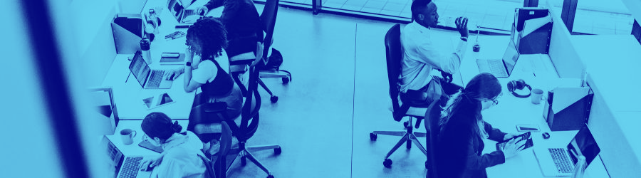 Varias personas trabajan con ordenadores portátiles y tabletas en una redacción de espacio abierto, sentadas en escritorios individuales, en tonos azules.