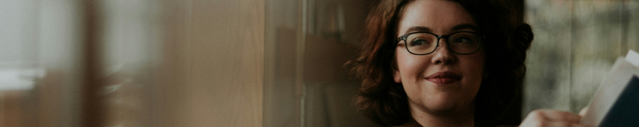 A woman with glasses and curly hair gently smiles while holding a book in a warm and cozy setting. A woman with glasses and curly hair gently smiles while holding a book in a warm and cozy setting.