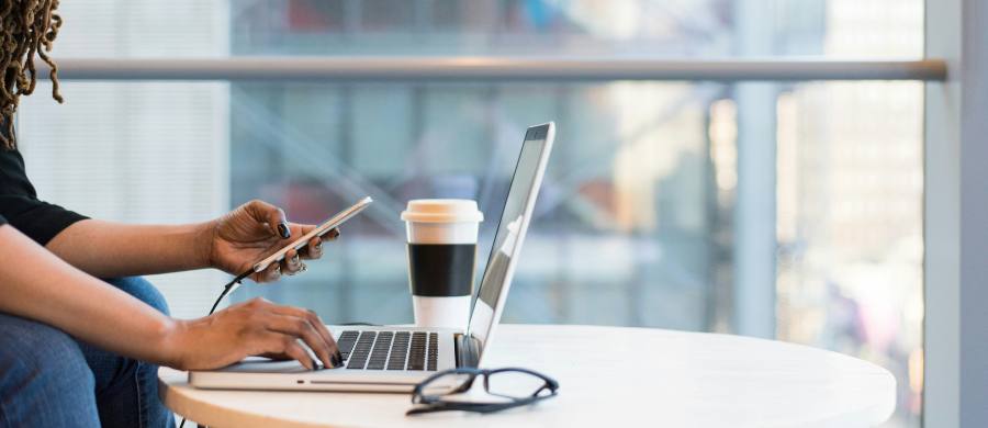 A person works on a laptop while holding a mobile phone in the other hand. On the table are some glasses and a takeaway coffee. The scene takes place in a bright space with large windows overlooking the city.