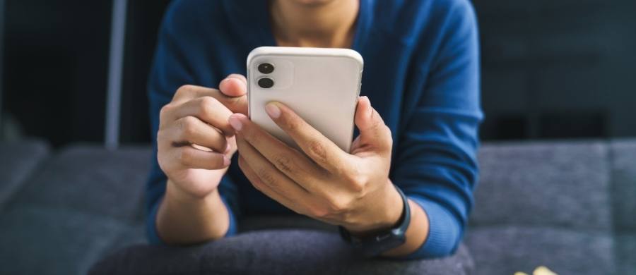 A person wearing a blue sweater holds a white phone while looking at the screen, sitting on a sofa in a cozy indoor setting.