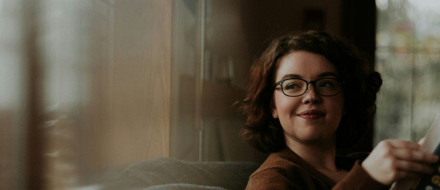 A woman with glasses and curly hair gently smiles while holding a book in a warm and cozy setting.