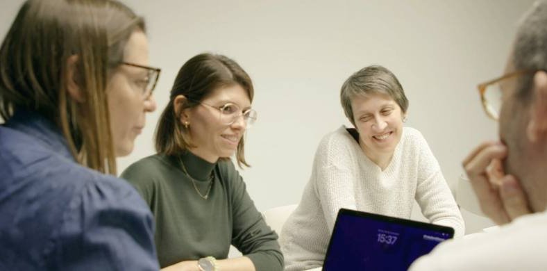 Captura del nou vídeo de la Biblioteca. Quatre persones en una reunió de treball, assegudes al voltant d’una taula. Dues dones amb ulleres escolten atentes, mentre una altra somriu. Un home amb ulleres reflexiona amb la mà a la barbeta.  Ambient distès i col·laboratiu.