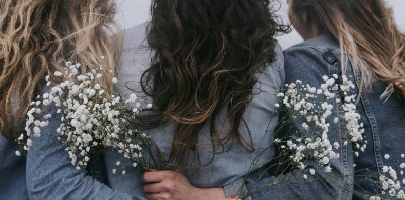 Tres mujeres de espaldas, con chaquetas vaqueras y el pelo suelto, se abrazan mientras sostienen ramos de flores blancas. La escena tiene un tono suave y melancólico.