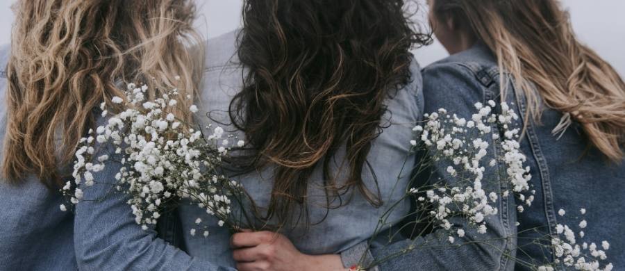 Tres mujeres de espaldas, con chaquetas vaqueras y el pelo suelto, se abrazan mientras sostienen ramos de flores blancas. La escena tiene un tono suave y melancólico.