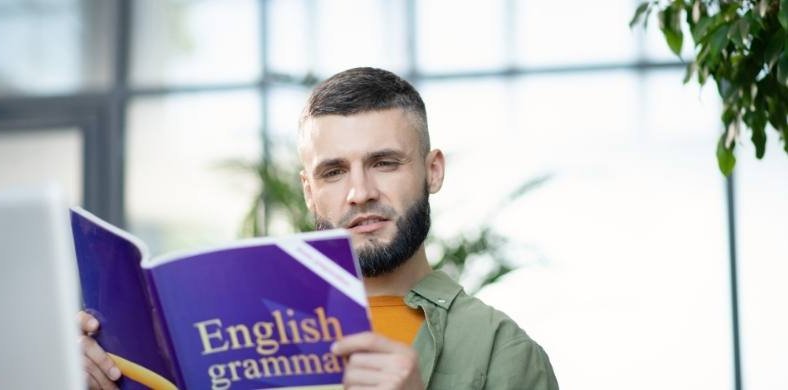 Un hombre joven con barba lee concentrado un libro de portada morada titulado “English grammar” en un espacio luminoso con plantas.
