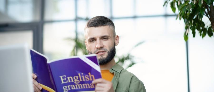 Un hombre joven con barba lee concentrado un libro de portada morada titulado “English grammar” en un espacio luminoso con plantas. Un hombre joven con barba lee concentrado un libro de portada morada titulado “English grammar” en un espacio luminoso con plantas.
