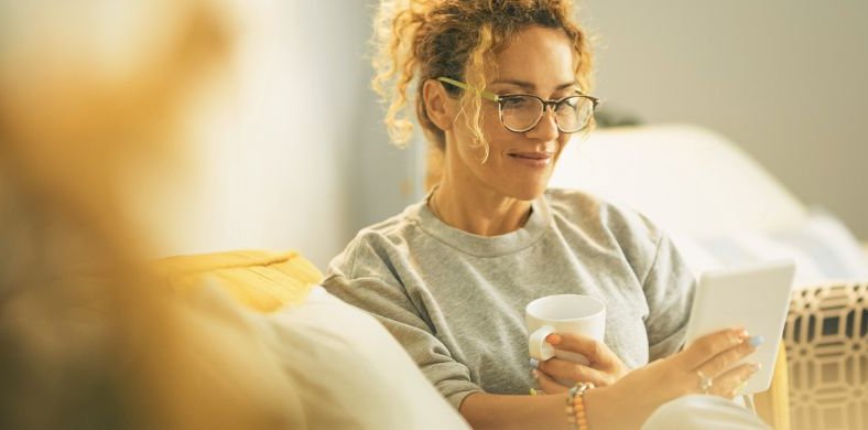 Una mujer sentada en el sofá leyendo un libro digital en la tableta con una taza en la mano.